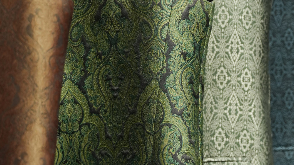 Close-up of a green and black ornate damask patterned fabric, with other patterned fabrics partially visible in the background.