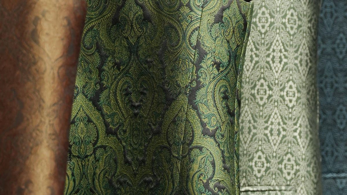 Close-up of a green and black ornate damask patterned fabric, with other patterned fabrics partially visible in the background.