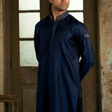A man wearing a dark navy blue long kurta with a textured patterned collar, standing in front of a rustic wooden door.