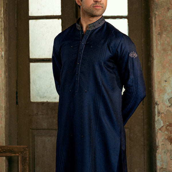 A man wearing a dark navy blue long kurta with a textured patterned collar, standing in front of a rustic wooden door.