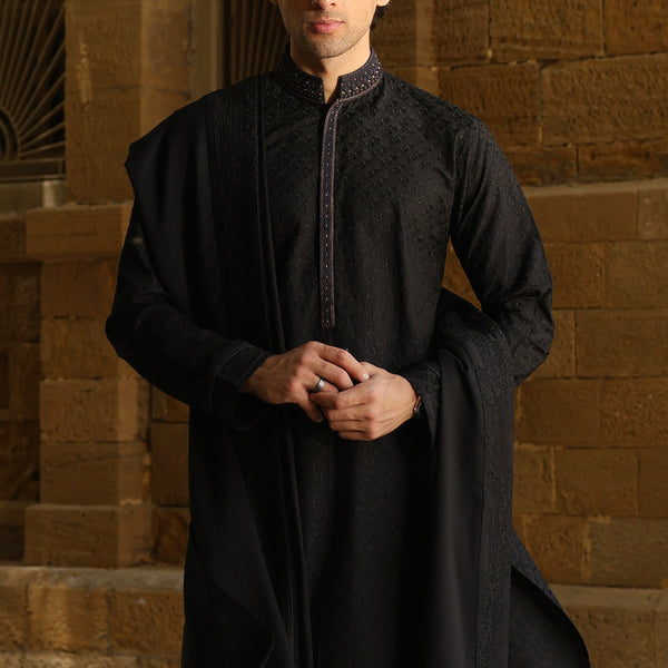 A man wearing a black embroidered traditional kurta with a matching draped shawl, standing in front of a rustic brown brick wall.