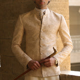 A man wearing a beige textured prince coat with a mandarin collar, holding a wooden cane, standing against a stone wall.