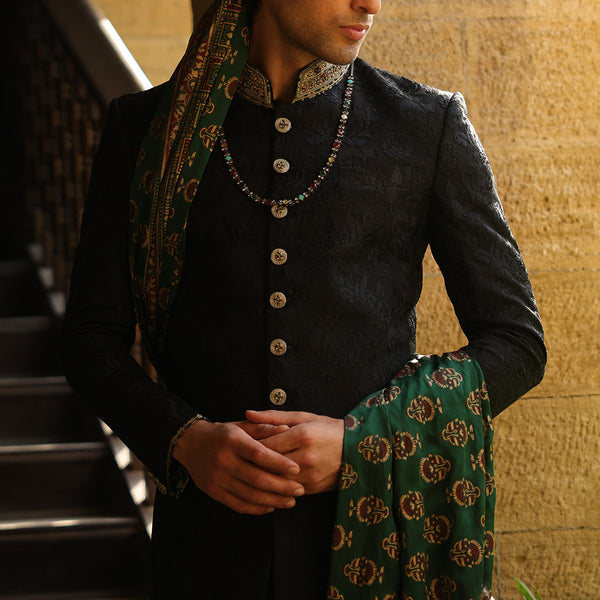 A man wearing a black textured sherwani jacket with ornate gold buttons and a green patterned dupatta draped over his shoulders, standing in front of a stone wall.