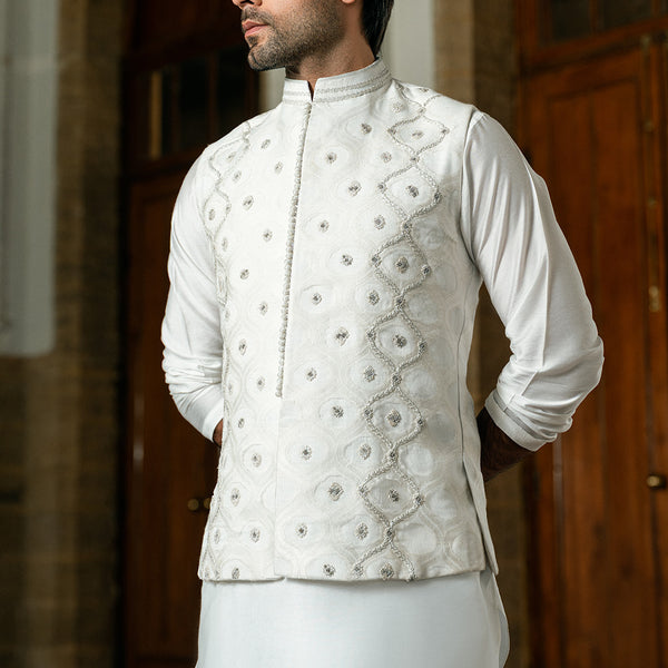 A man wearing a white long-sleeve kurta with a matching embroidered vest, photographed indoors against a blurred wooden door background.